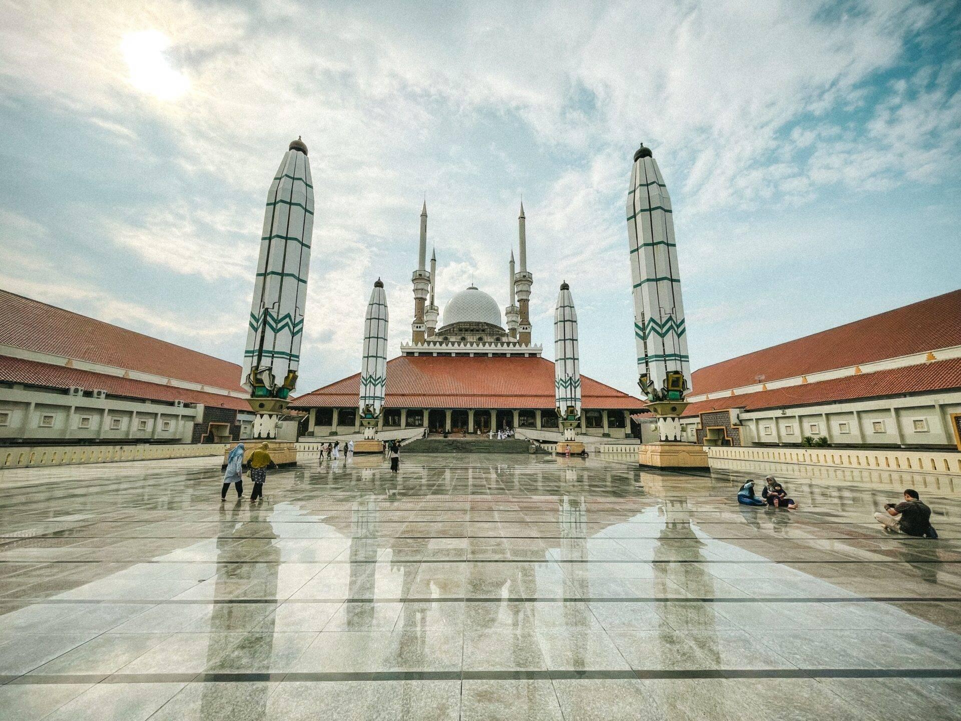 A grand mosque with a white dome and ornamental minarets, a reflective paved courtyard and majestic Islamic architecture