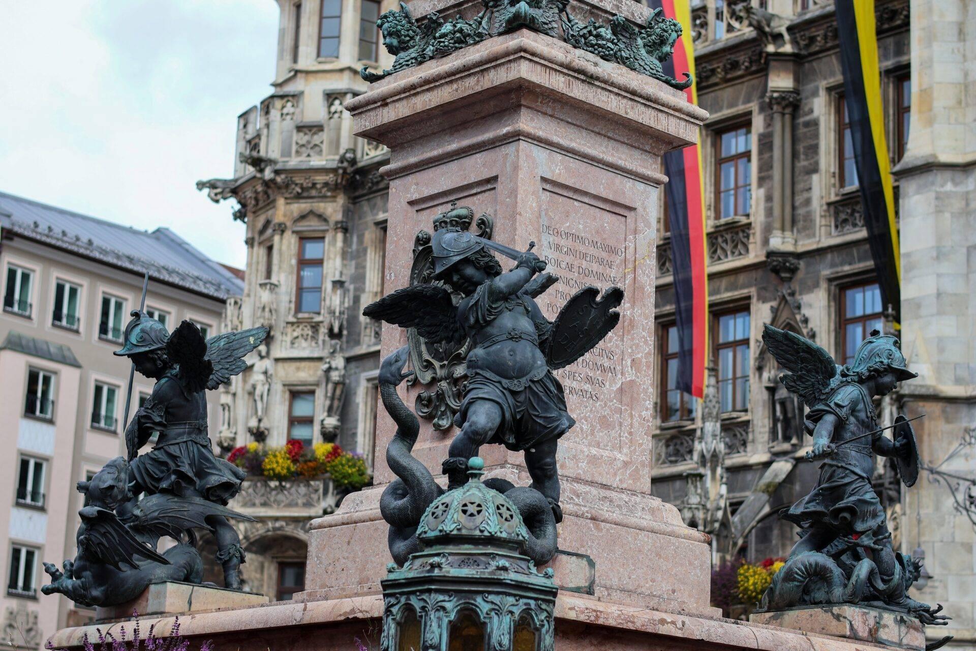 Monument with winged bronze angel statues on a stone plinth in a historic European square surrounded by classical architecture