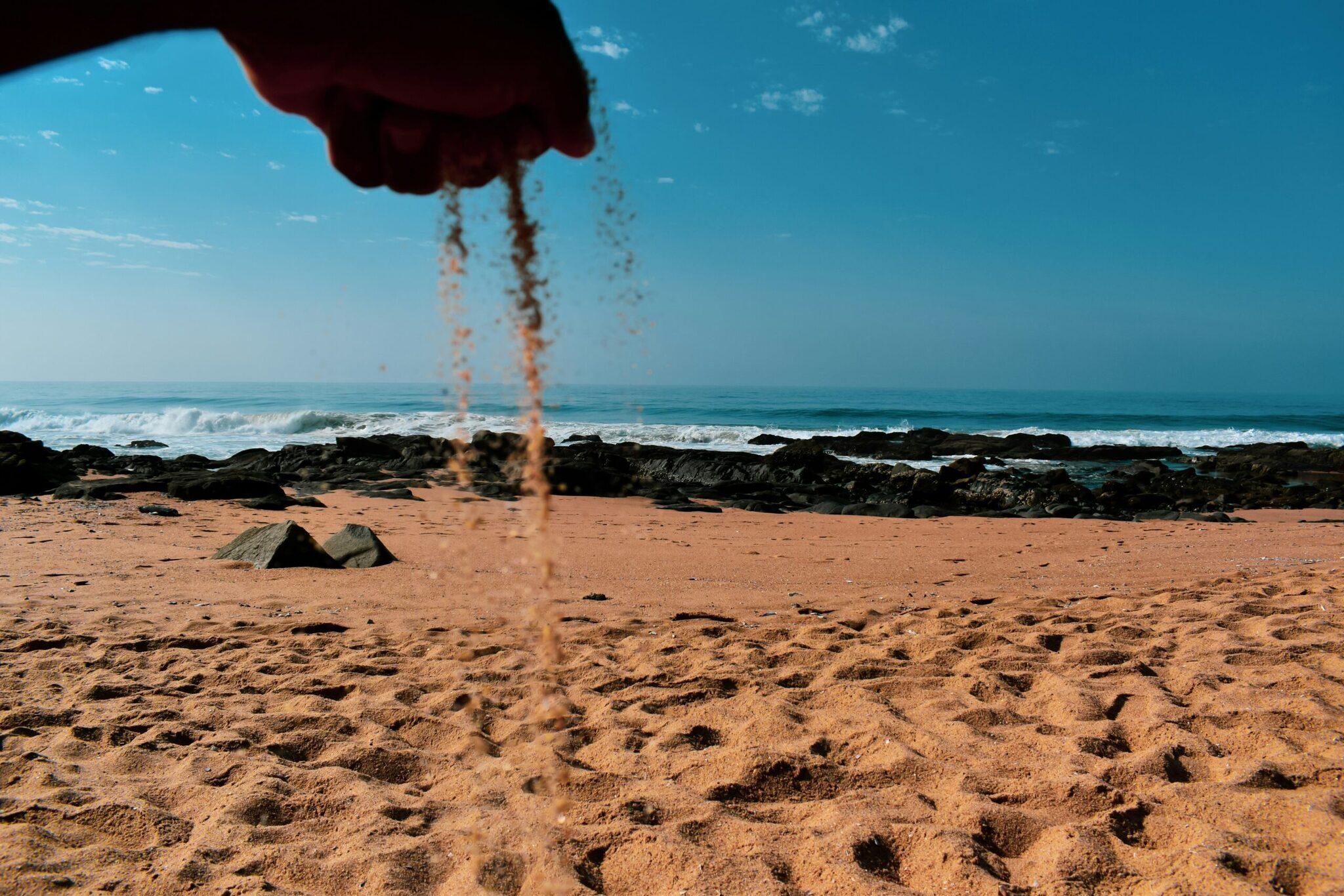 A hand pouring fine sand on a rocky beach with a view of the ocean and waves