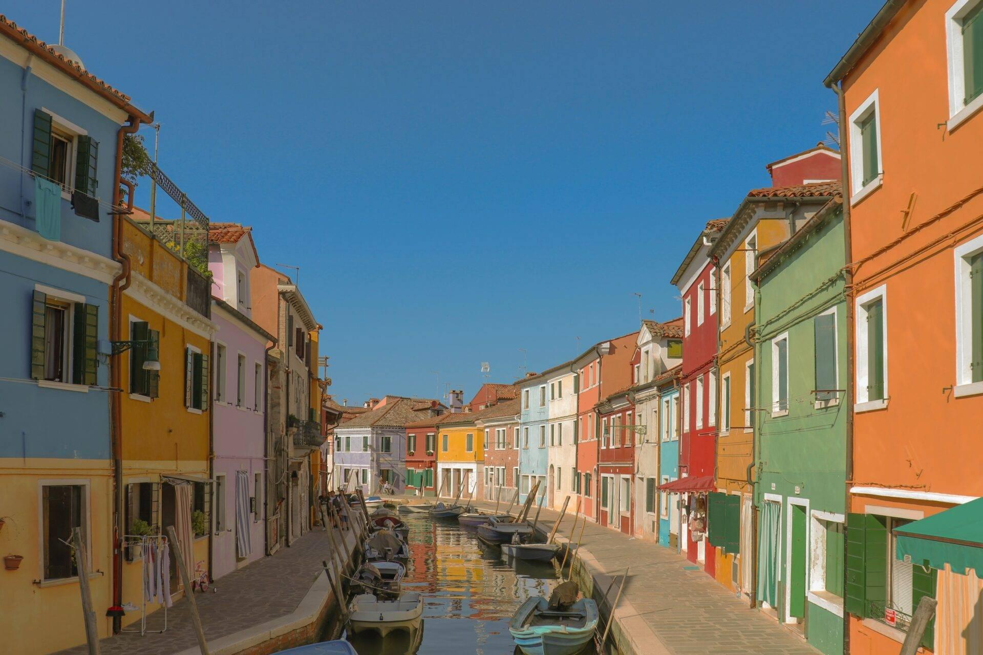 A colourful Venetian alleyway lined with yellow, orange and green houses, with boats moored along the canal